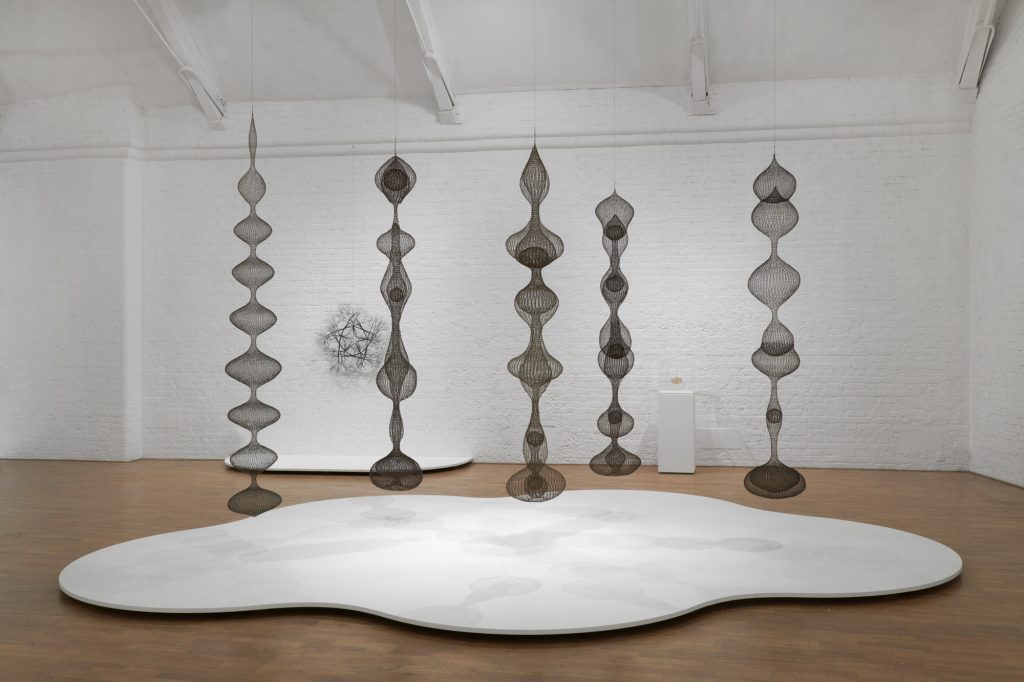 Five sculptures hanging from a high ceiling. They are made of looped wire and are in abstract bulbous shapes. A white flat abstract form is on the floor beneath them. A wire sculpture hangs on the wall and a small plinth holds a small gold wire sculpture behind them.