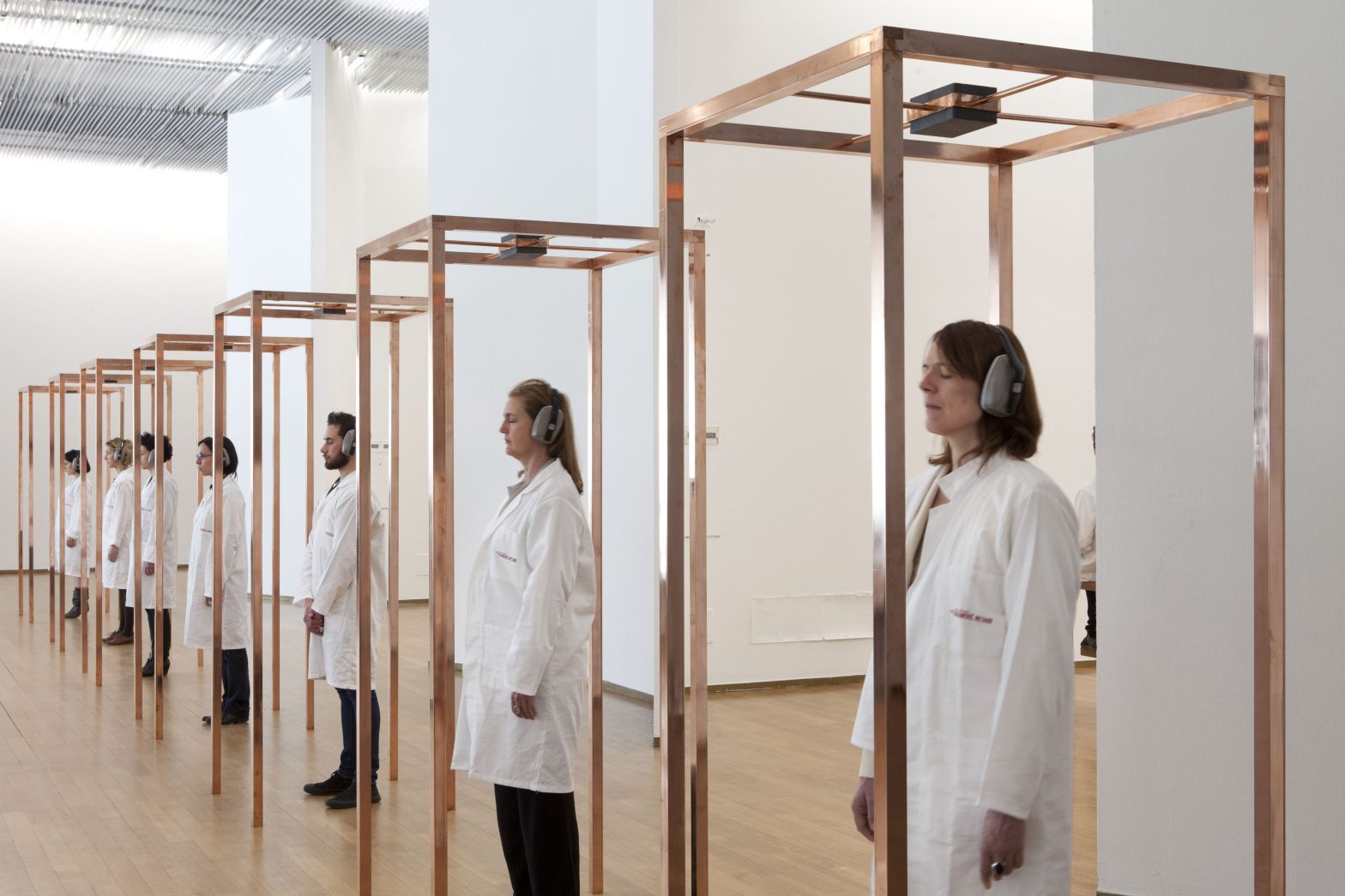 A line of tall rectangular copper structures in a row inside an art gallery. A line of people wearing lab coats and headphones are each standing in the centre of each structure, facing outwards.