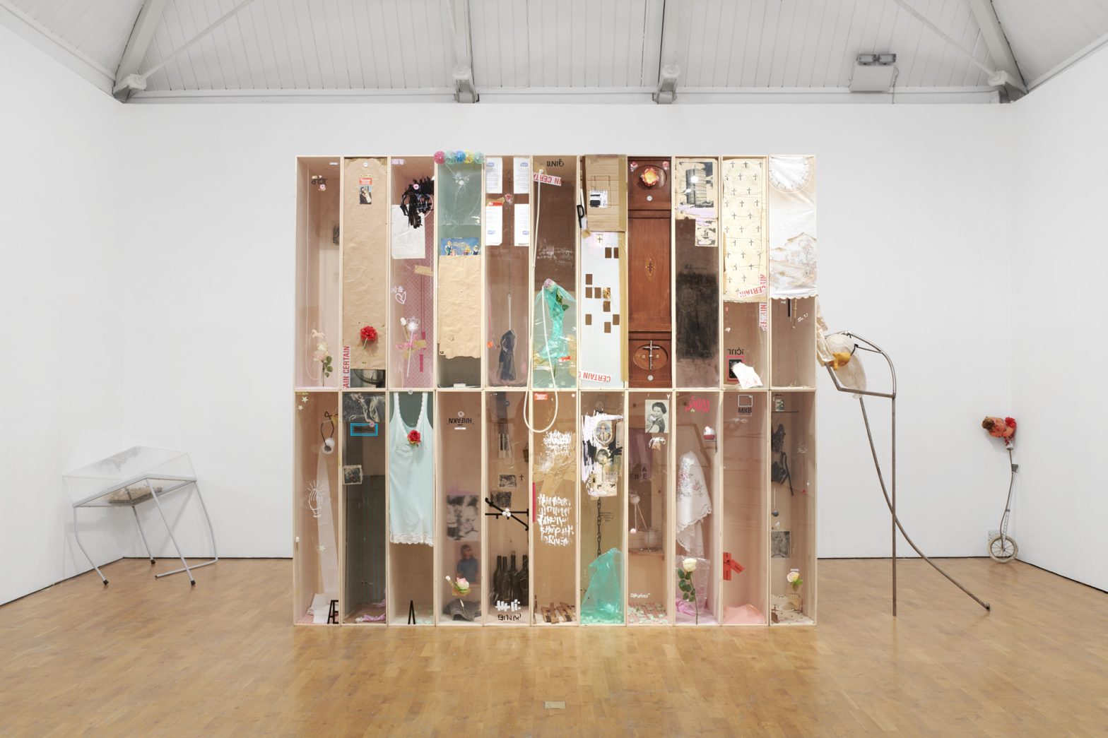 An art installation made up of rows of open wooden boxes filled with a variety of objects, images, clothes and jewellery. On either side of the sculpture are artworks made of metal rods and wheels.