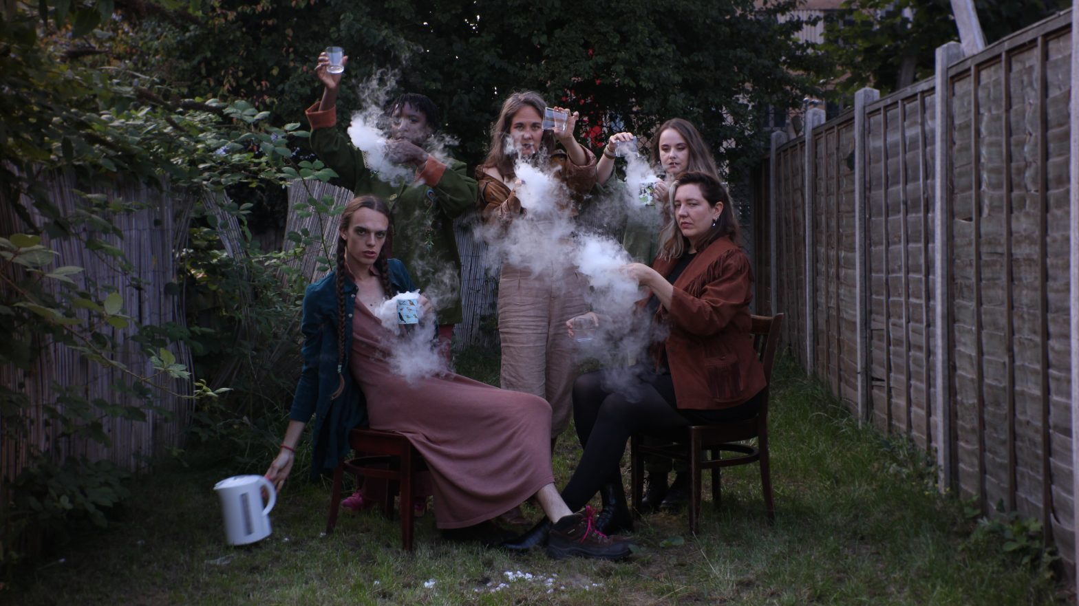 Group of 5 people sitting in a garden holding steaming teacups and a kettle.