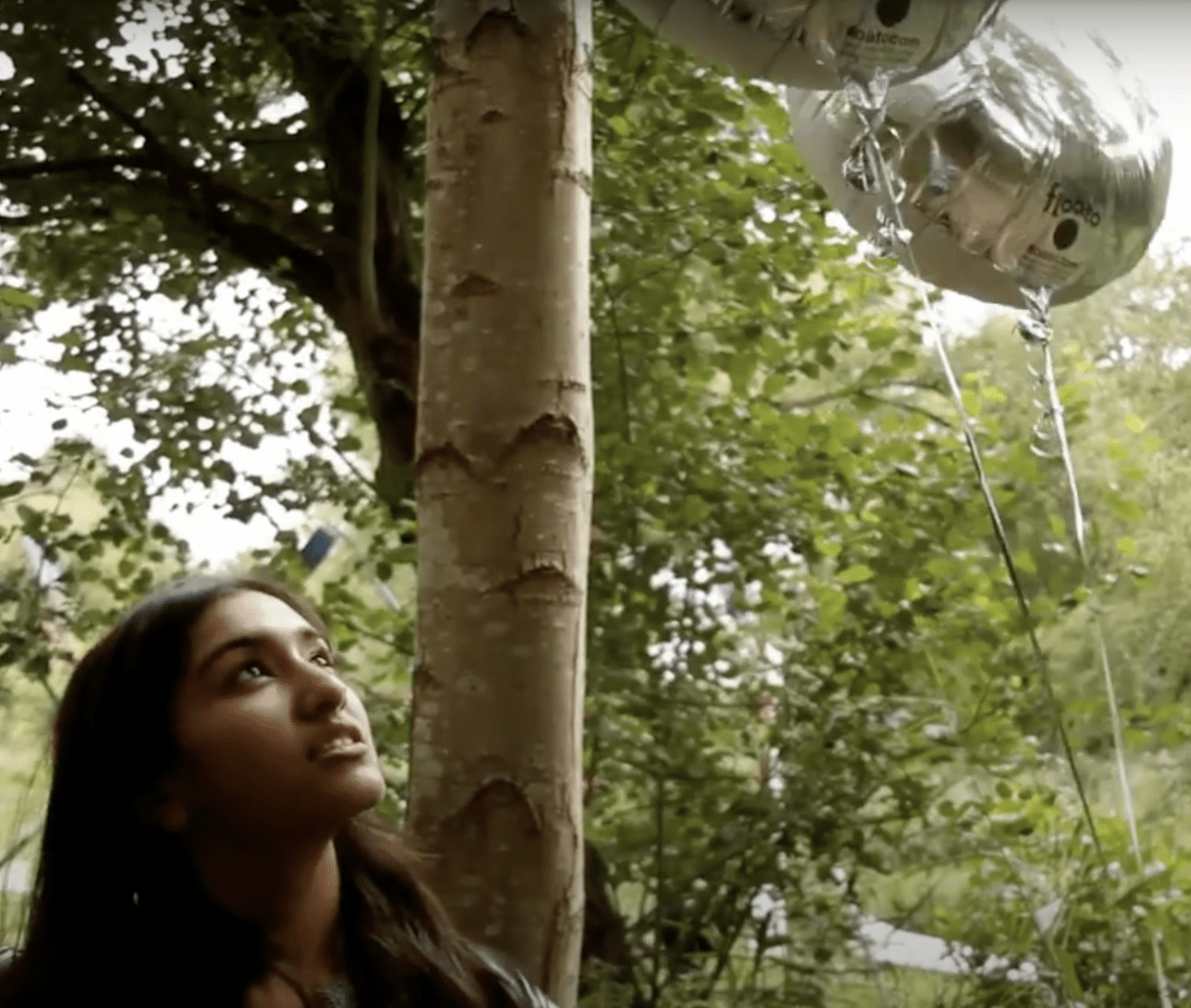 Young woman amongst trees looking up two silver balloons.