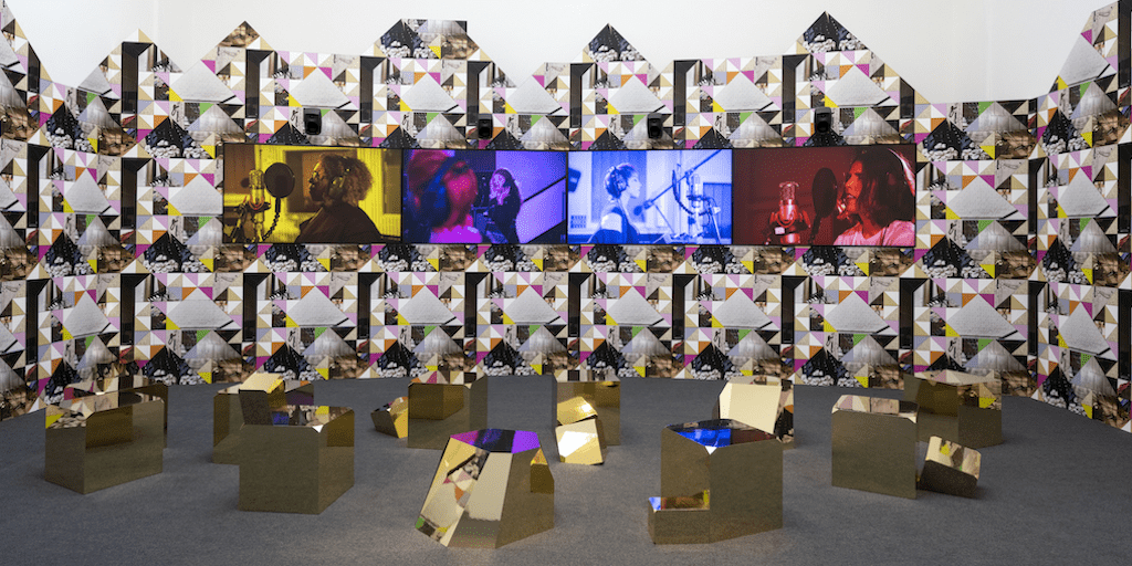 Four large flat screens with singers against a wall surrounded by a kaleidoscope of images, colourful shapes and shiny gold geometric sculptures of different shapes on a grey floor.