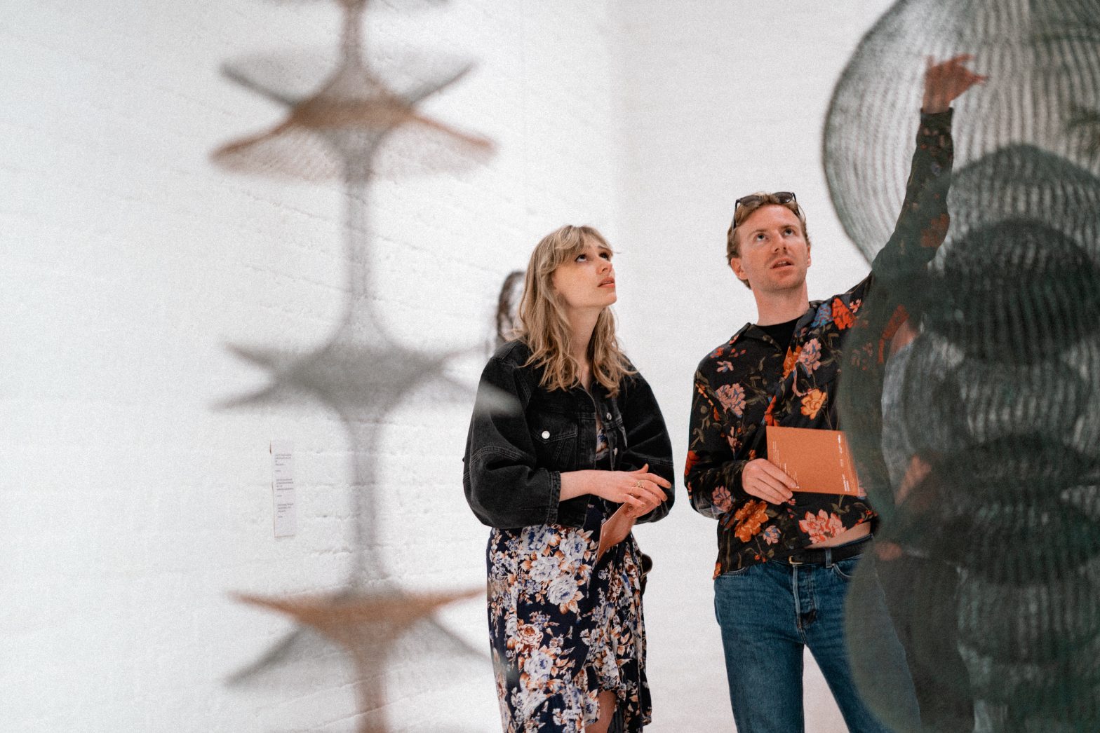 Two people standing in front of a wire hanging sculpture by Ruth Asawa. Another hanging sculpture is visible but blurred in the foreground.