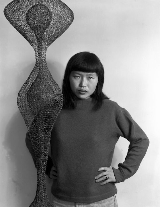 Young woman with dark hair and a heavy fringe stands with arms on her hips, in front of a large rounded hanging sculpture made of looped wire. The woman wears a dark jumper and stares directly.