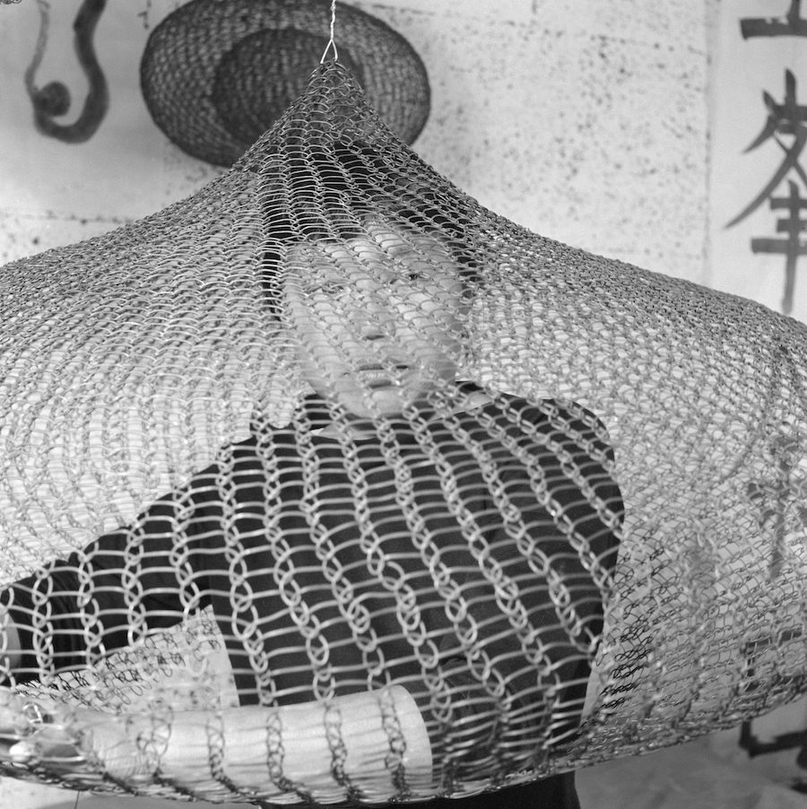 A young woman with dark hair and a fringe behind a large looped-wire sculpture which covers her face. She wears a dark long sleeved black top and in the background are hand painted Japanese words and another wire sculpture.