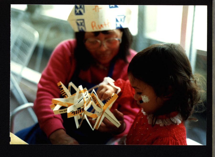 A woman wears a paper hat and shows a young child wearing faceprint a star shaped folded paper sculpture.