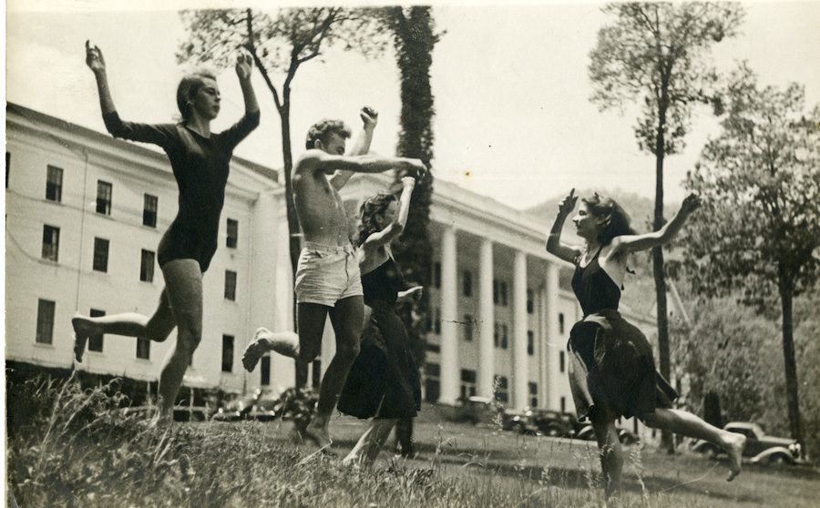 Four young people dance in the grass with arms and legs making expressive gestures, in front of a large building with columns and many windows.