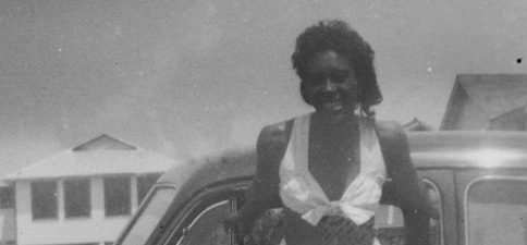 Black and white photo of a young Black woman (Alma Stone Williams) wearing a white cropped top and shorts leaning against a car smiling.