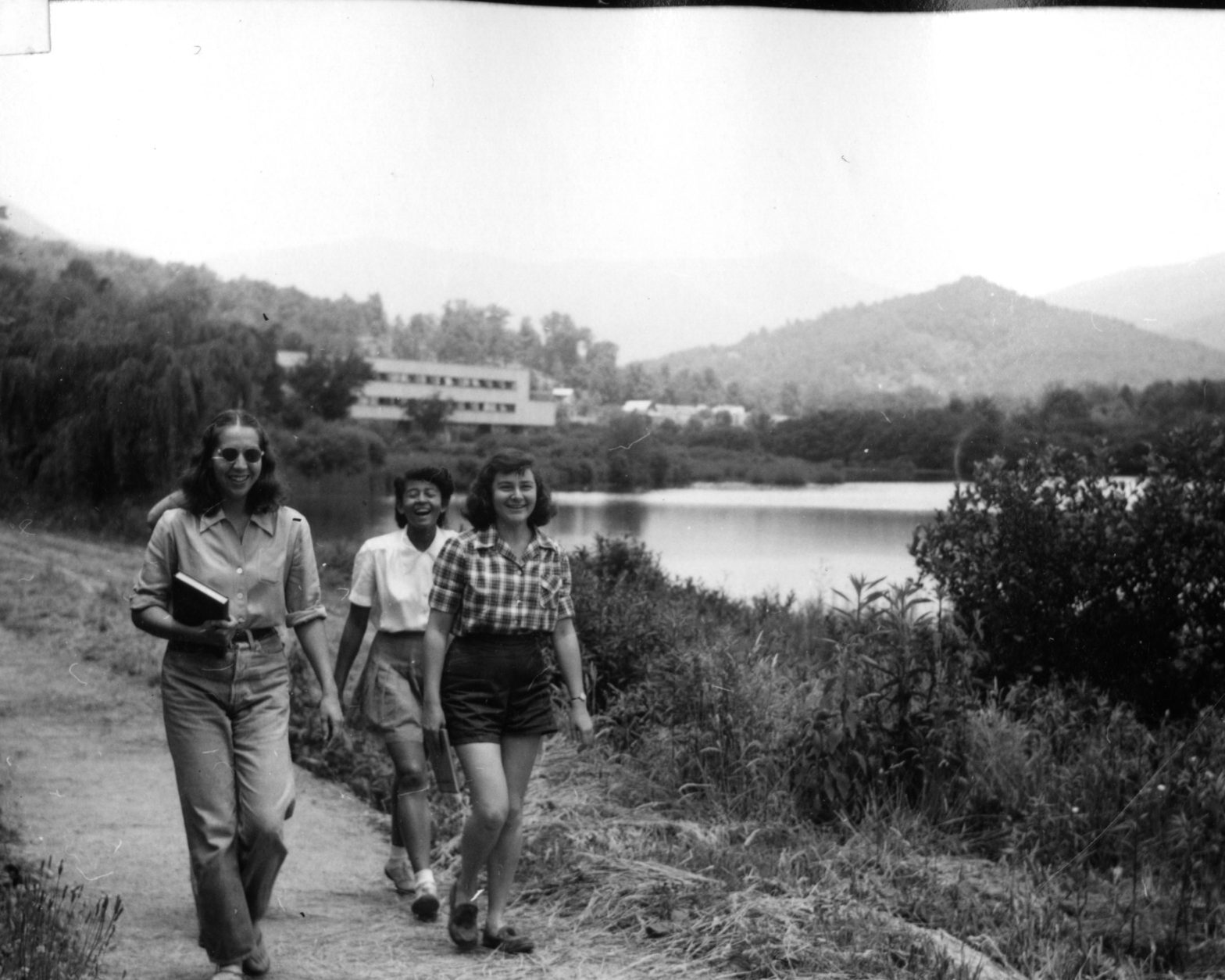 Black and white photo of three laughing young women, walking along a country path next to a large lake, with mountains in the background. There is also a white modernist building in the background.