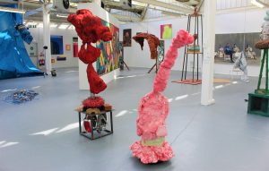 Two brightly coloured bulbous sculptures in a brightly lit gallery space. They seem to be made of metal car parts and are coated in bulbous red and pink paint.