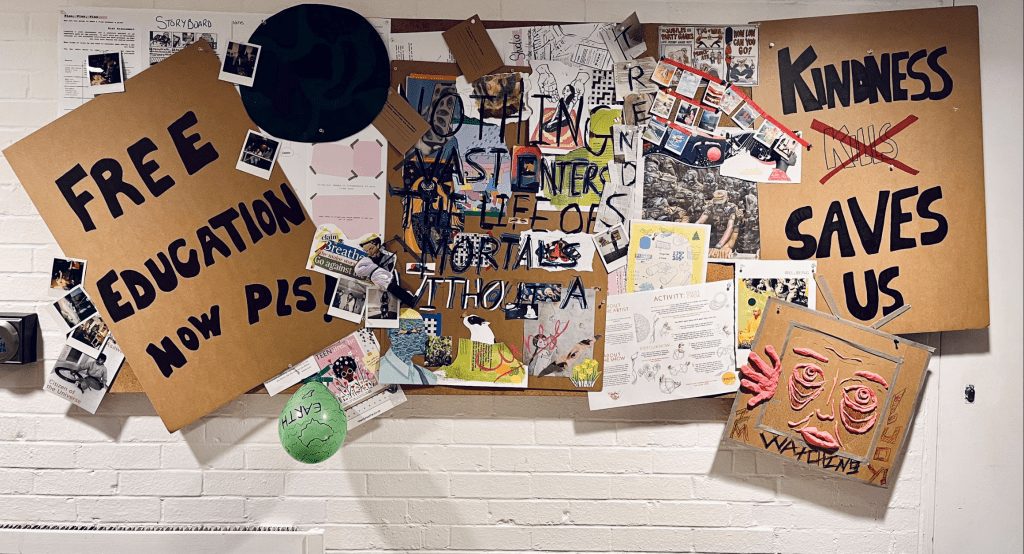 Cork board on a white wall covered in handwritten signs, drawings and newspaper clippings. The big billboards read: 'Free education now pls' and 'kindness saves us'