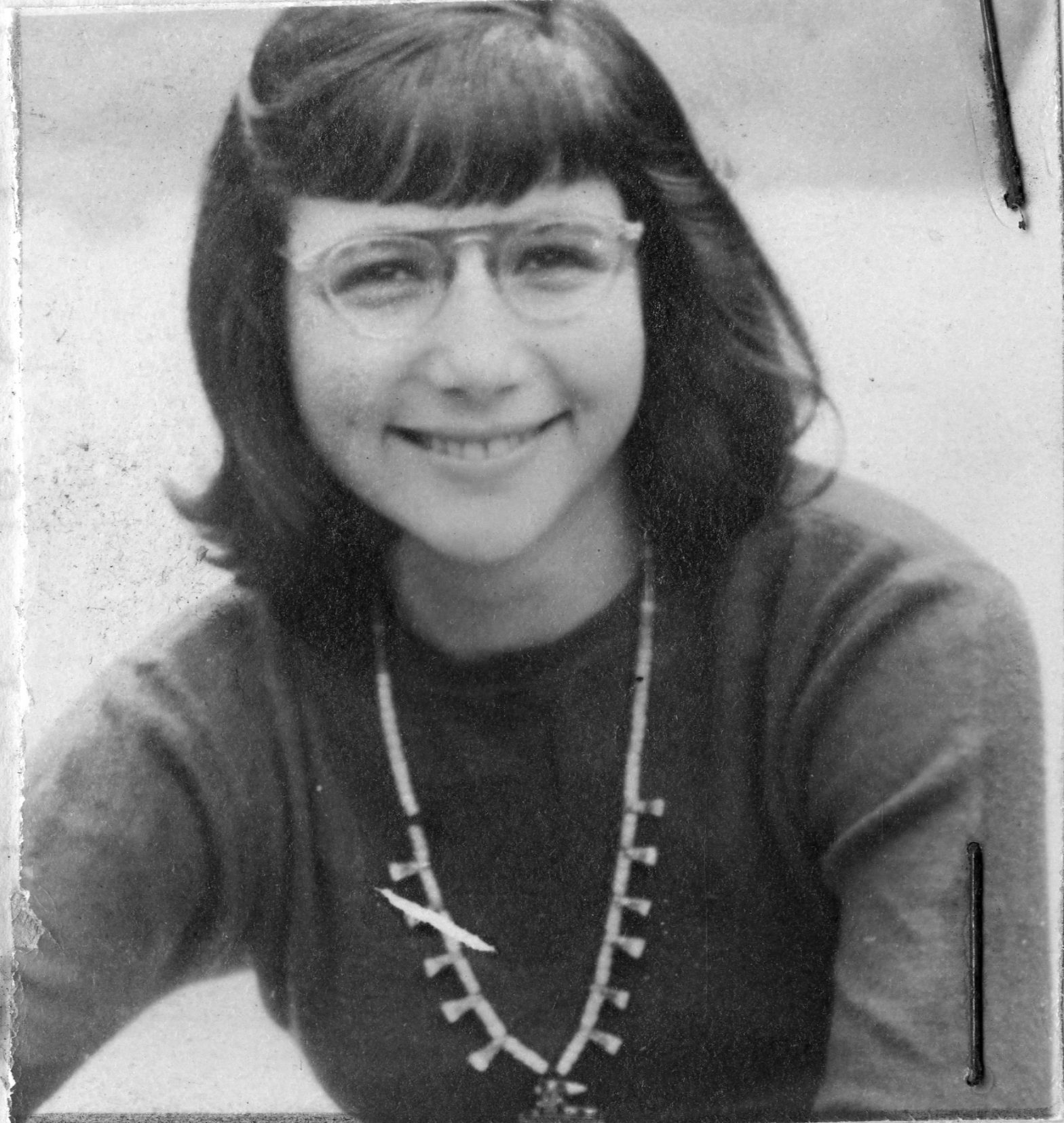 Black and white photo of a white woman with short straight hair and a fringe, wearing glasses and smiling at the camera.