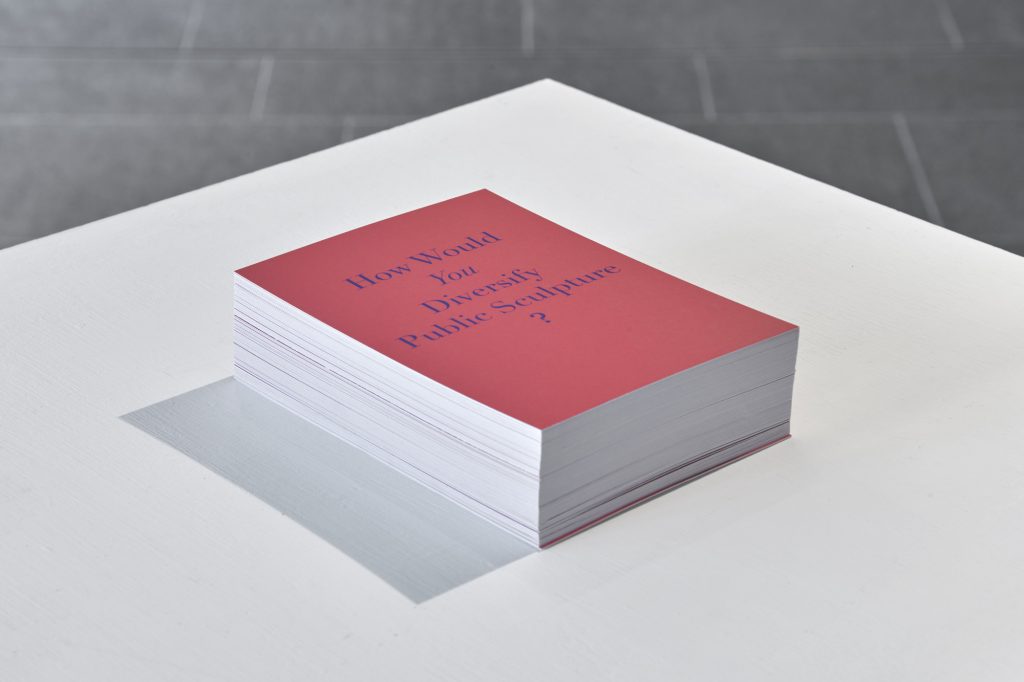 A stack of red postcards with the words 'How would you diversify public sculpture?' on them, on a white surface