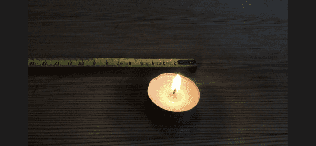 Photograph taken from above of a tea-light candle burning on a wooden surface, with an extended tape measure next to it, measuring the tea-light at 5 centimetres.