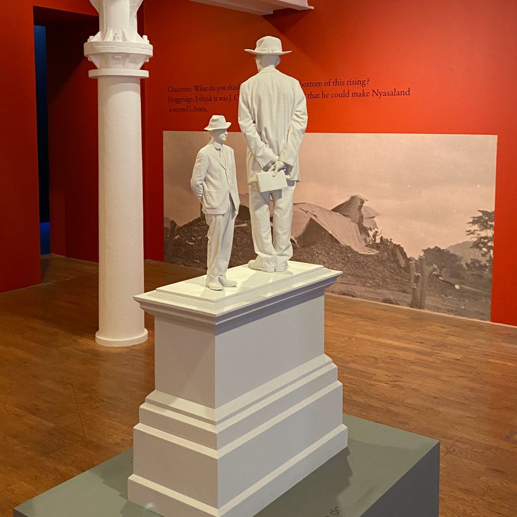 A sculpture of two men, one much larger than the other, both wearing hats, standing on a plinth. Behind on a red wall is a black and white photograph showing a building destroyed as a pile of rubble.