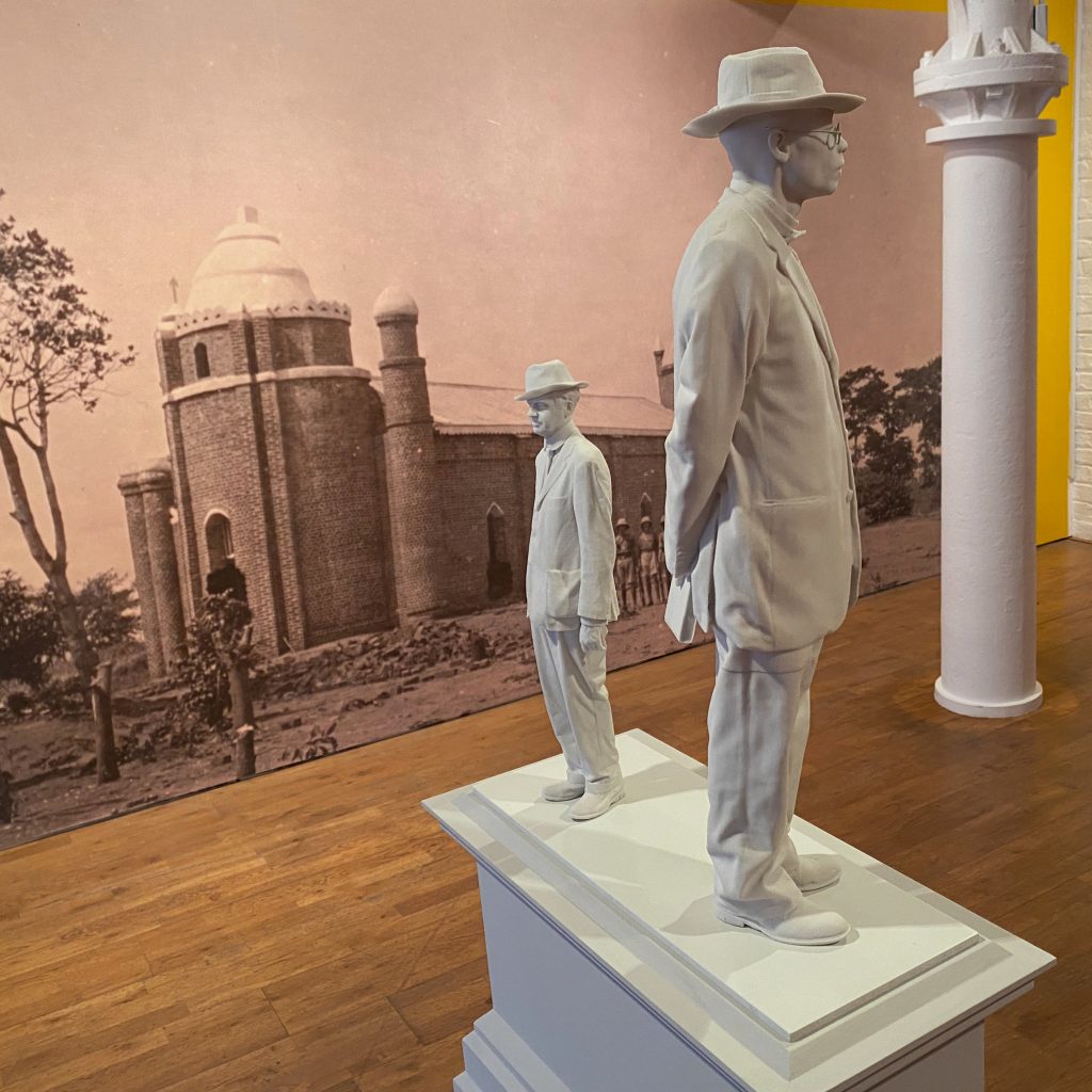A sculpture of two men, one much larger than the other, both wearing hats, standing on a plinth.Behind on a yellow wall is a black and white photograph showing a church with armed soldiers in front of it