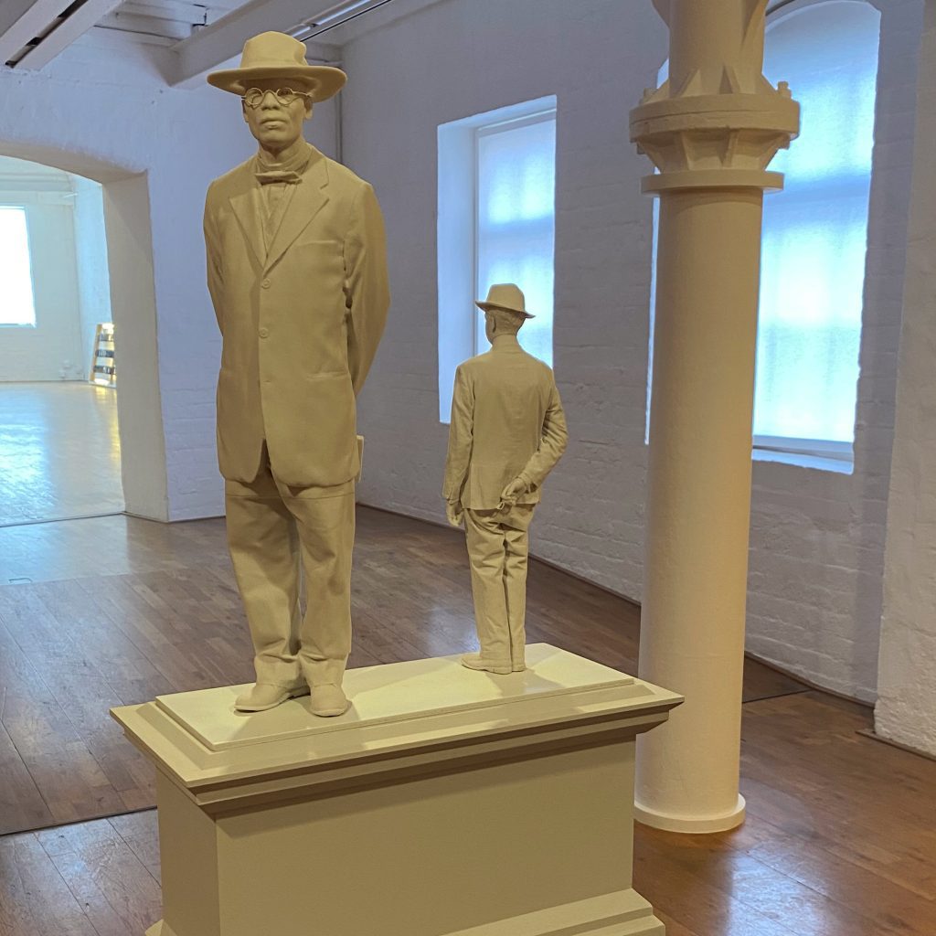 A sculpture of two men, one much larger than the other, both wearing hats, standing on a plinth.