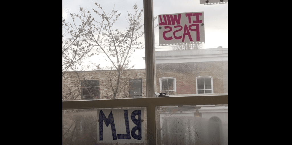 Photograph of a window taken from indoors. The view through the window is a residential street, with a tall tree. There are two signs up in the window facing outwards, one reads "BLM" and one reads "IT WILL PASS"