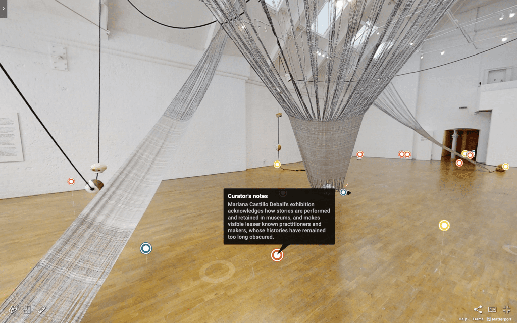 Screenshot of the online virtual tour of Mariana Castillo Deball's exhibition at Modern Art Oxford called Between making and knowing something. Large swathes of woven fabric hang from the ceiling in the Upper Gallery of Modern Art Oxford. There are small coloured tags across the virtual space. One of them is open and reads 'Curator's Notes: Mariana Castillo Deball’s exhibition acknowledges how stories are performed and retained in museums, and makes visible lesser known practitioners and makers, whose histories have remained too long obscured."