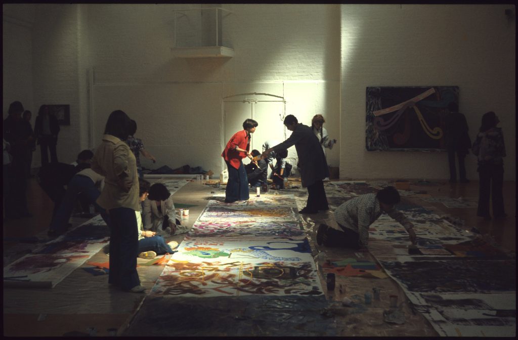 Several people standing and crouched over large abstract paintings, in a large gallery space with sunlight filtering through.