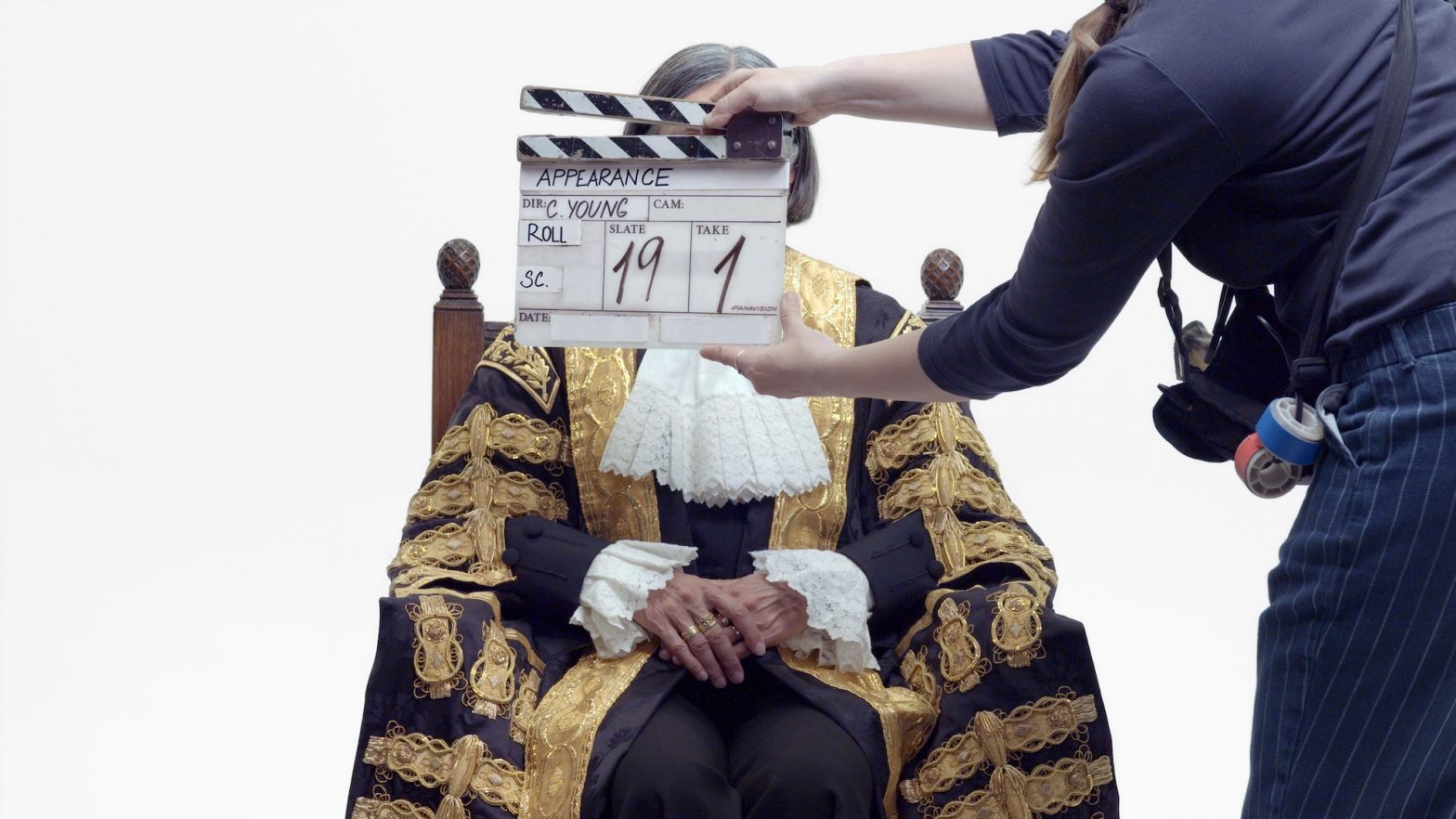 A film clapperboard being held in front of a person in an ornate gold and black costume, who is sitting with their hands in front of them against a white background.