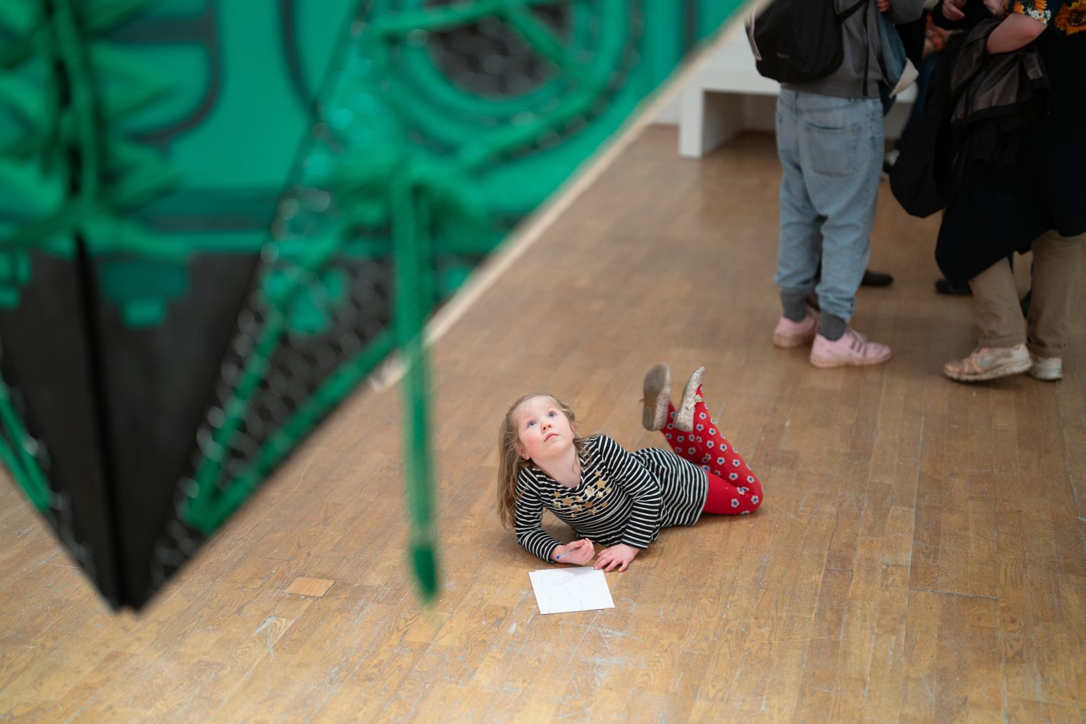 A young girl wearing a stripey top and brightly coloured tights lies on the floor with a pen and paper, looking up at a huge green sculpture.