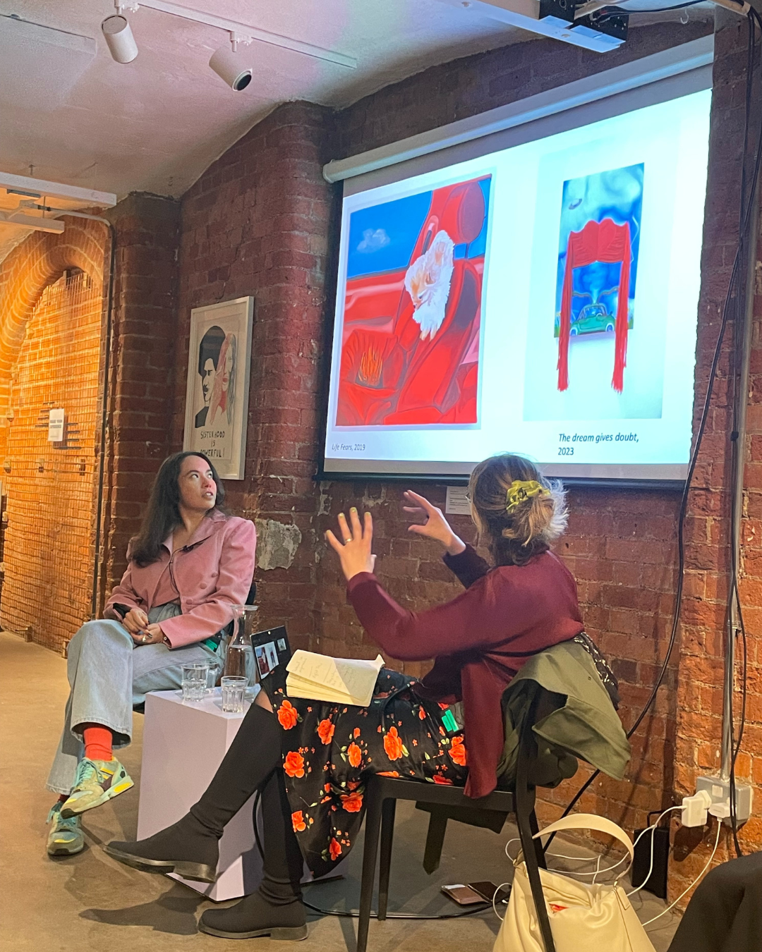 A photo showing artist Frieda Toranzo Jaeger who has long brown hair wearing a pink top and blue jeans in conversation with Dr. Larne Abse Gogarty who is wearing a red jumper and black skirt.