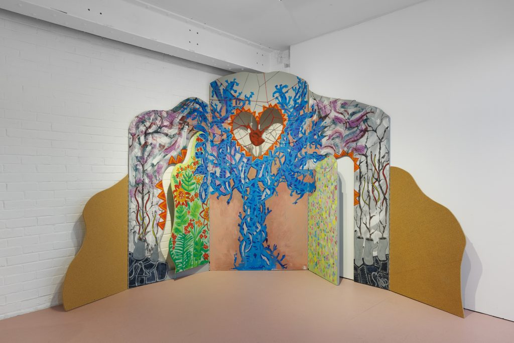 A photo of a collaborative  artwork created by Modern Art Oxford's Young Creatives. It is a multicoloured, multi-panelled arch showing a tree with plants growing. Inside the tree is a heart. Embroidery runs throughout the piece. 