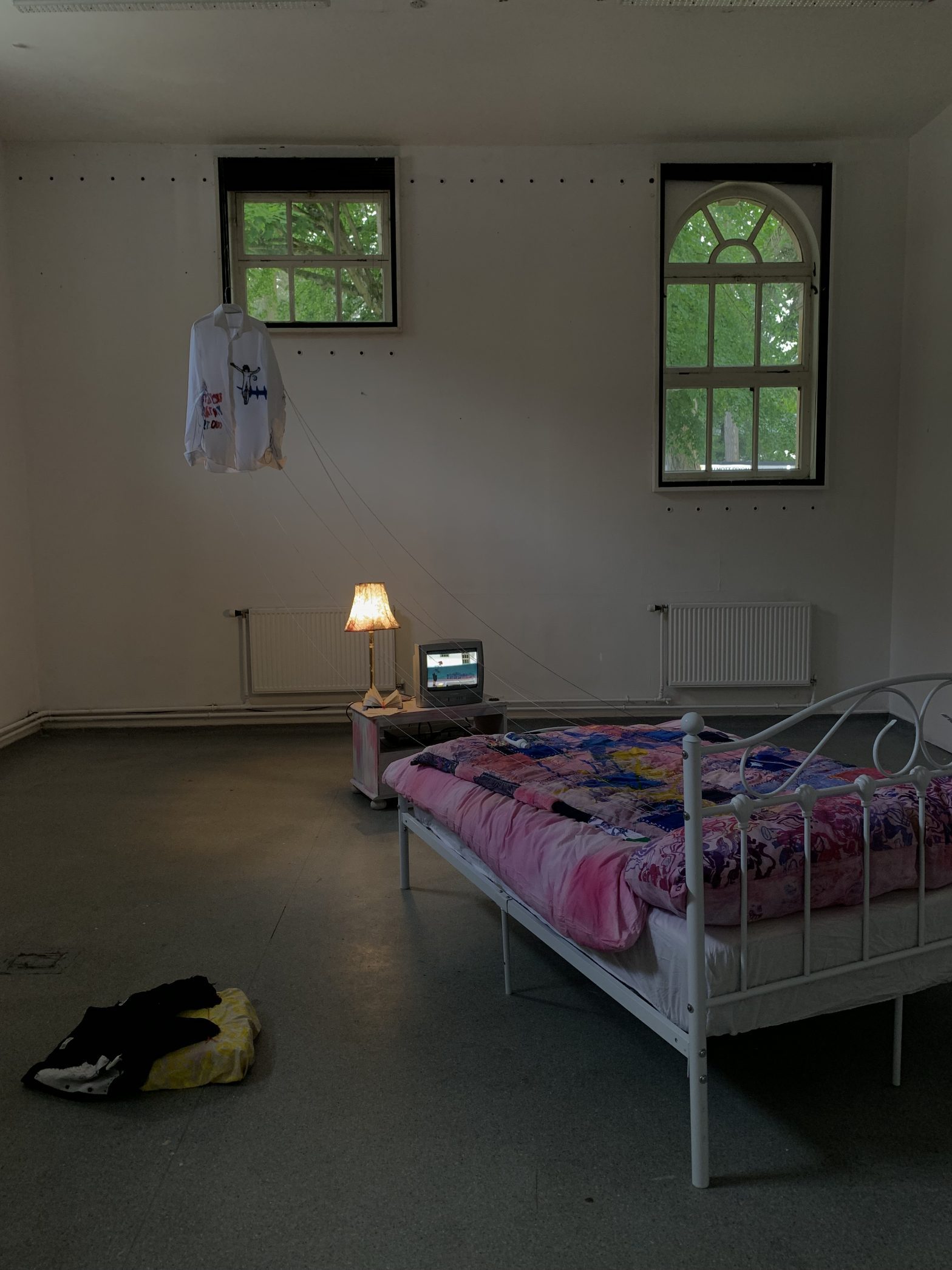 A photograph of an installation. It shows a bed covered in a hand embroidered quilt facing a table with a lamp and small tv on it.