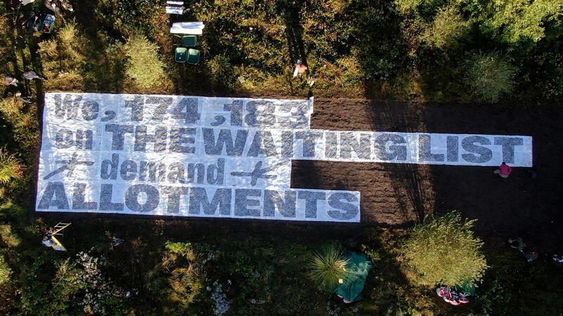 An aerial photograph showing a huge banner reading: 'We, 174,183 on THE WAITING LIST *demand* ALLOTMENTS. It sits on privately owned but unused land which could become common ground.