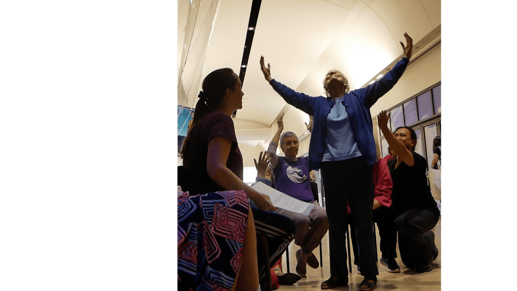 An elderly woman stands in the centre of a circle of people with her arms raised above her head.