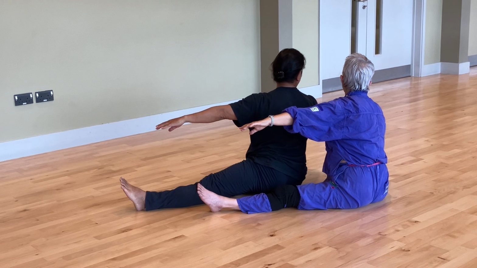 Two elderly women sit on the floor together, with their arms and legs outstretched in dance.