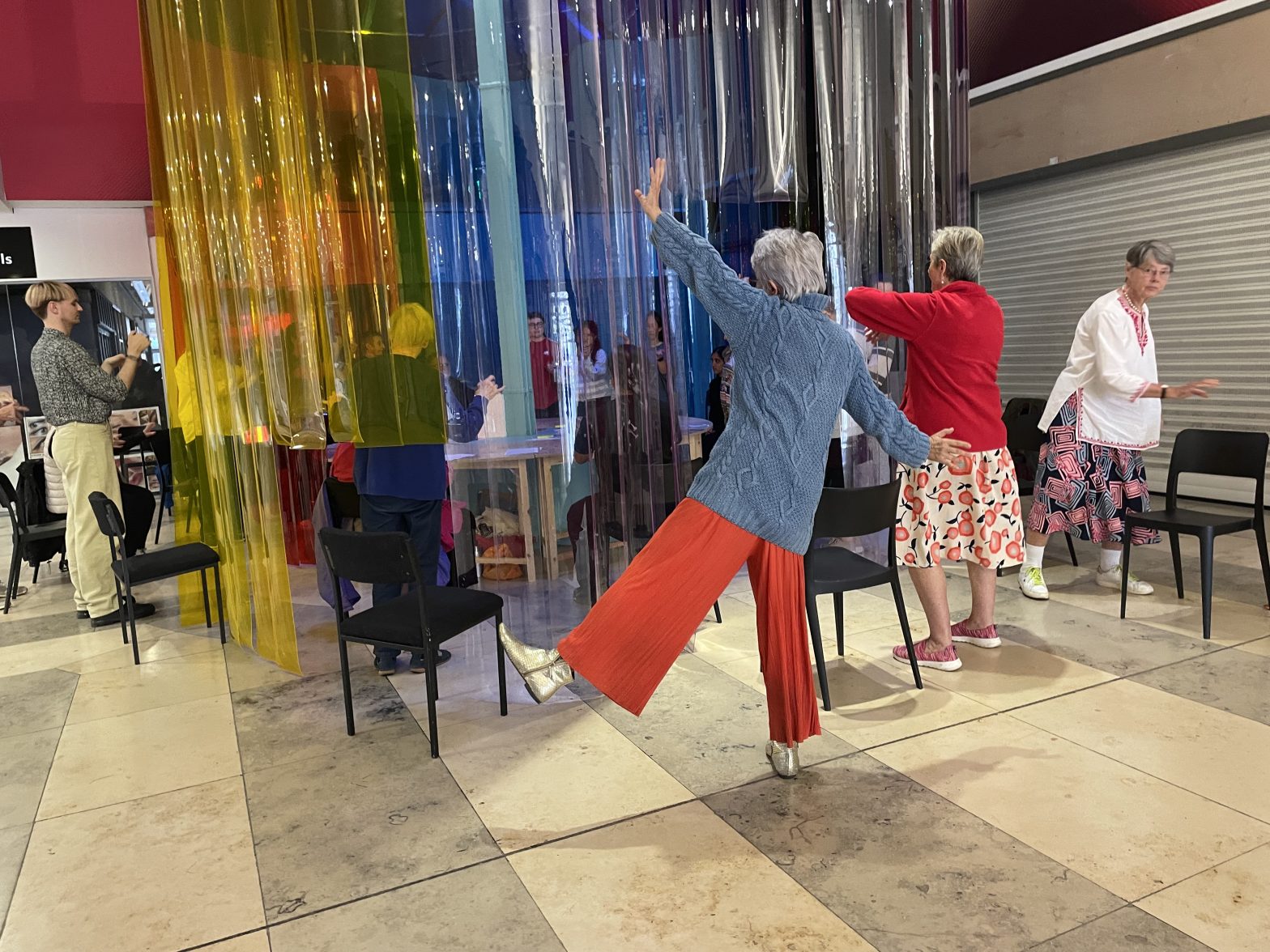 A photograph of a group of people, ranging in ages dancing around a brightly coloured canopy of hanging plastic sheets.