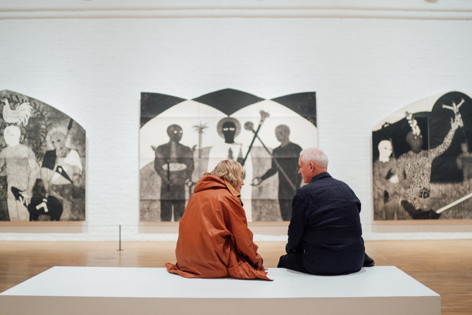 An elderly couple sit on a bench in a brightly-lit gallery.