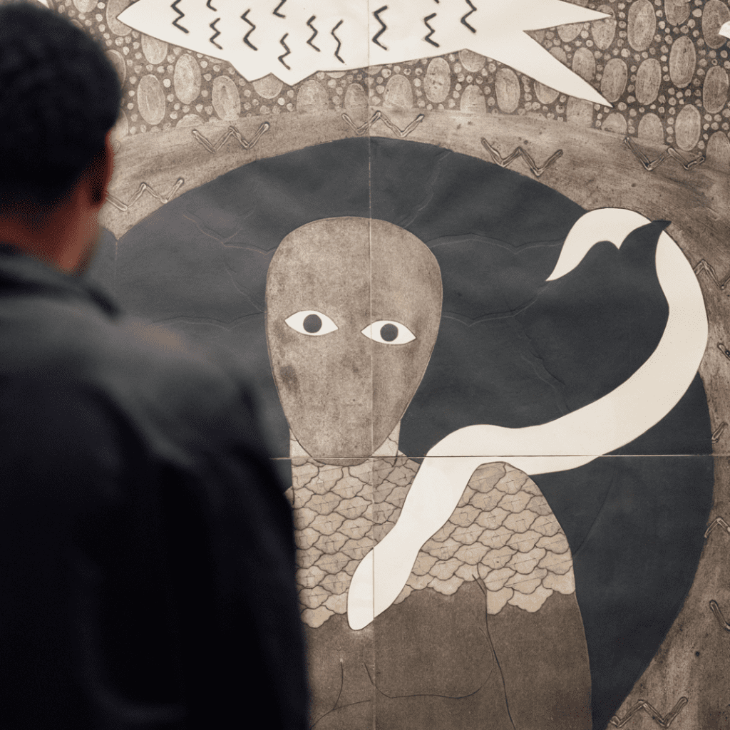 A photo of a man in an art gallery looking at a collograph print by artist Belkis Ayón. The print depicts a figure with no eyes or mouth staring directly into the camera. A snake is wrapped over the shoulder and a fish is floating above their head. 