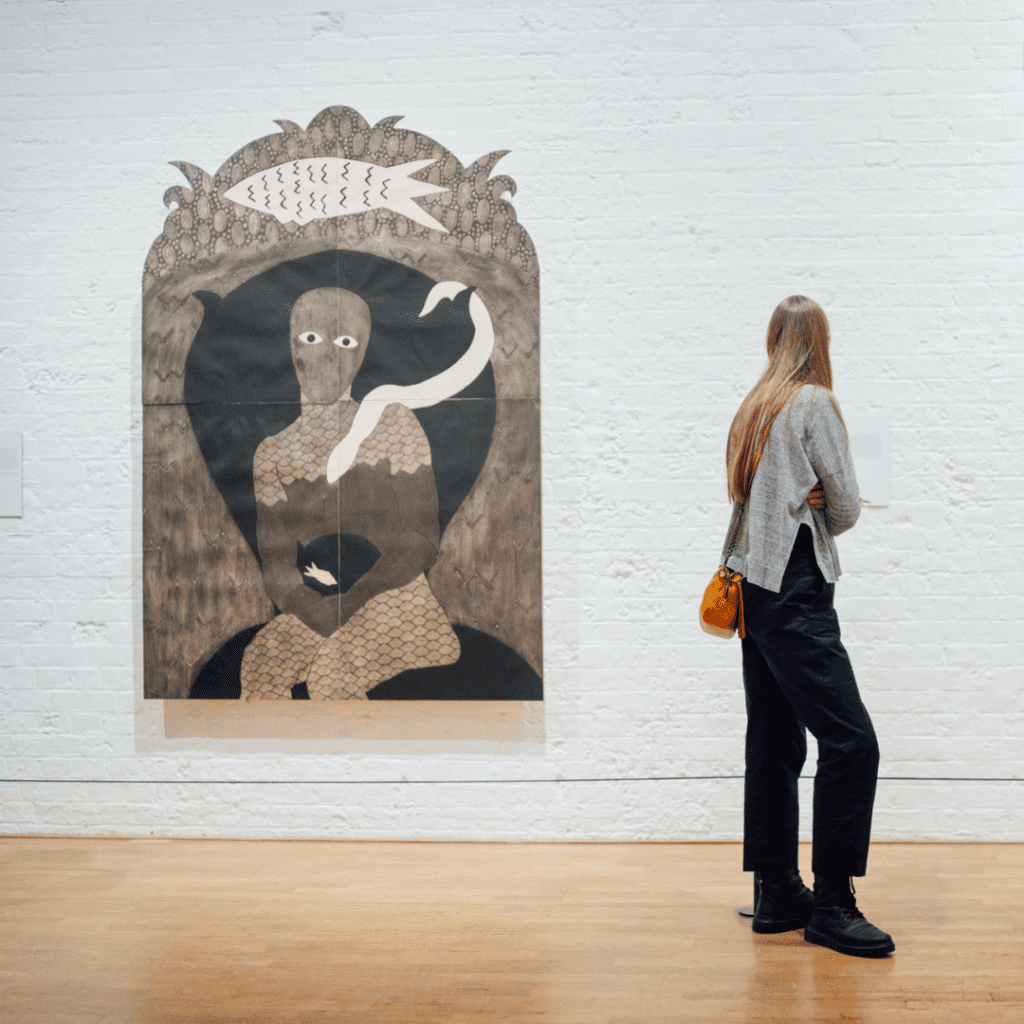 A photo of a woman with long brown hair, grey jumper and black jeans standing in front of a print by Belkis Ayon in a gallery