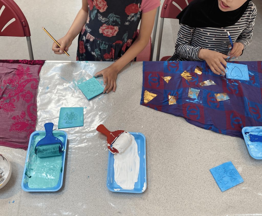 Two children use paint rollers and foam to print shapes onto fabric.